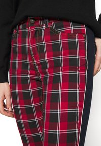 Gros plan sur un pantalon à carreaux rouge et noir avec des coutures blanches et jaunes, porté avec un haut noir, montrant la poche latérale et la main posée à proximité.