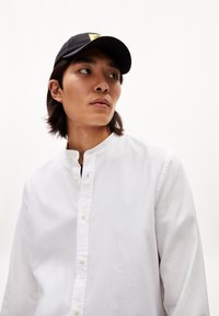 Witte blouse met staande kraag, voorzien van een kleine geborduurde accent. Zwarte pet met een geel logo.