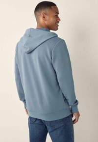 Sudadera con capucha de textura azul y corte relajado. Cuenta con un bolsillo tipo canguro y puños de canalé. Combinada con jeans de mezclilla.