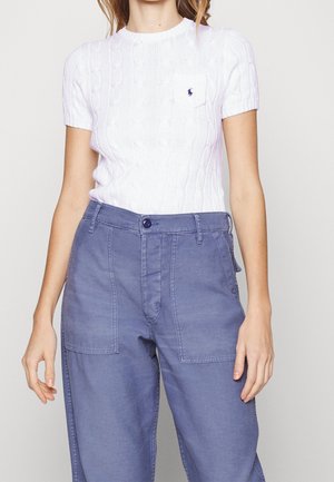 Vrouw draagt een witte, korte mouw, gebreide top met een klein borstzakje en blauwe hoge taillebroek met grote voorzakken.