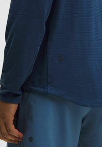 Langärmliges, blaues Sporthemd mit strukturiertem Stoff und einem runden Logo am Saum, kombiniert mit hellblauen Shorts.