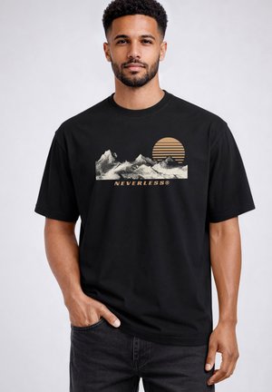 Man wearing black t-shirt with beige mountain range and sun graphic above the word "NEVERLESS®," standing with one hand in pocket.