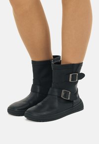 Botas de tobillo de cuero negro con un acabado texturizado, cierre lateral con cremallera y correas decorativas con hebillas plateadas. Suela de goma para mayor tracción.