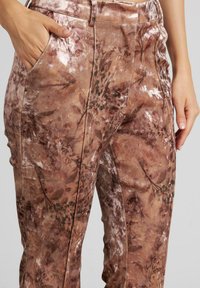 Braune Samthose mit einem floralen Muster in gedämpften Farbtönen. Sie besitzen Seitentaschen und eine glatte, glänzende Textur.