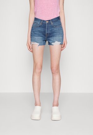 Jeans-Shorts in Mittelblau mit fransigen Säumen und verwaschenen Details, kombiniert mit weißen Slippern. Das Model steht vor einem neutralen Hintergrund.
