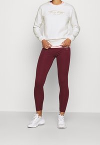 Felpa bianca con logo dorato stampato, indossata sopra un top rosa, accompagnata da leggings bordeaux. Presenta scarpe da ginnastica bianche.