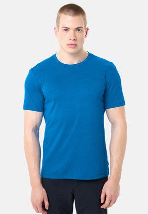 Junger Mann mit kurzen Haaren, der ein eng anliegendes blaues T-Shirt mit kurzen Ärmeln und schwarze Hosen trägt, vor einem schlichten hellen Hintergrund steht.