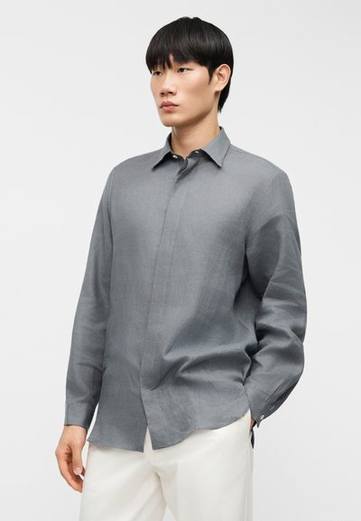 Γκρι λινό πουκάμισο με μυτερό γιακά, μακριά μανίκια και κούμπωμα με κουμπιά μπροστά. Απαλή υφή με διακριτικά σχέδια. Συνδυασμένο με λευκό παντελόνι.