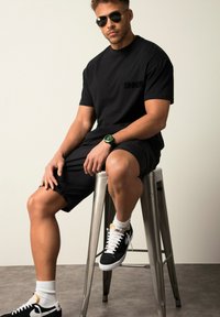 Schwarzes Baumwoll-T-Shirt mit "SINNER"-Logo, kombiniert mit schwarzen Shorts. Trägt eine grüne Uhr, schwarze Sneaker und weiße Socken, sitzt auf einem Metallhocker.