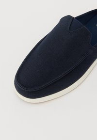Marine slip-on sko i strukturert stoff med lav profil. Sålen er hvit, og det er subtile stikninger langs kantene.
