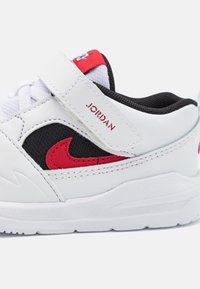 Bílé tenisky s černou síťovanou jazykem, červenými akcenty po straně a na pásku, vyrobené z kůže a gumy. Mají suchý zip a texturovanou podrážku.