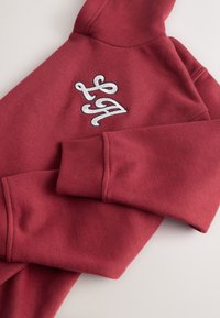 Felpa rossa in tessuto morbido, con logo ricamato in bianco. Dotata di cappuccio con coulisse e polsini a coste.