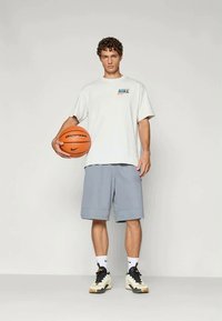 Valge Nike t-särk värvilise logoga, hallid lühikesed püksid ja mustad/valged tossud. Hoides oranži korvpalli, mille peal on tekst "playground".