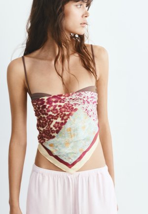Giovane donna che indossa un top foulard multicolore con motivi floreali e astratti sopra spalline di reggiseno marrone e pantaloni rosa chiaro, guarda verso destra.