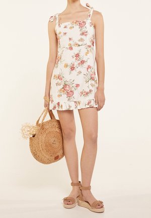 Femme portant une robe blanche à fleurs avec des liens aux épaules, tenant un sac rond tissé avec des fleurs séchées, et portant des sandales compensées beiges à bride à la cheville.