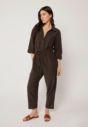 Femme aux longs cheveux foncés portant une combinaison marron foncé boutonnée avec manches retroussées et des sandales plates marron, debout devant un fond uni.