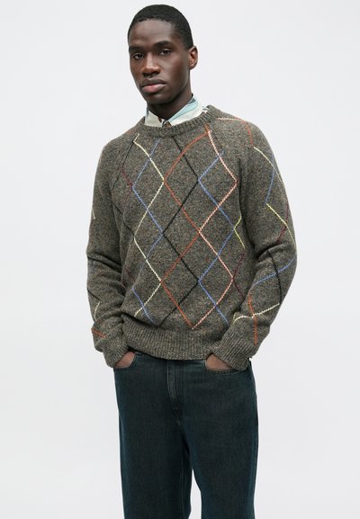 Barna texturált pulóver színes argyle mintával, bordázott mandzsettákkal és szegéllyel, inget alatta viselve, sötét farmerral párosítva.