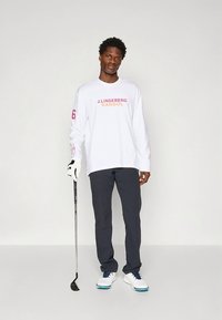 Valge pika varrukaga särk punase ja roosa logotekstiga, ühendatud tumedate pükste ja valgete vabaajajalatsitega. Hoides golfikeppi ja kandes kindaid.