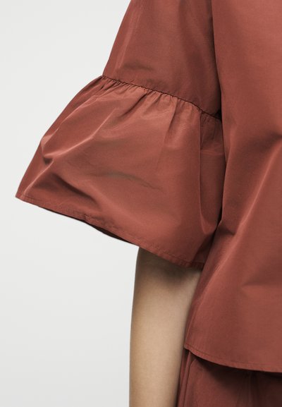 Λεπτομέρεια μανικιού σε ρούχο χρώματος σκουριάς με σχεδιασμό με σούρα και φαρδύ άνοιγμα πάνω από γυμνό χέρι σε απλό φόντο.