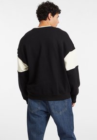 Schwarzes Sweatshirt mit cremefarbenen Akzenten an den Ärmeln, Rundhalsausschnitt, lockere Passform und geripptem Saum. Kombiniert mit hellblauen Jeans.