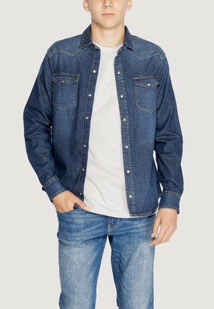 Camicia di denim blu scuro con chiusura a bottoni, due tasche sul petto e cuciture a contrasto. Indossata sopra una maglietta bianca e abbinata a jeans blu.