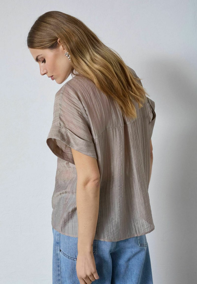 Femme aux longs cheveux châtain clair portant un chemisier ample taupe à manches courtes et un jean bleu clair sur un fond uni.