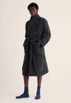 Homme portant un peignoir sombre à rayures verticales et des chaussettes bleues, debout devant un fond clair uni.