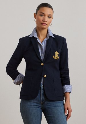 Kvinne iført en marineblå blazer med gullknapper og emblem over en blå og hvitstripet skjorte, kombinert med blå jeans og gylne hoops-øredobber.