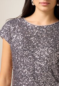 Srebrna cekinowa bluzka z okrągłym dekoltem i krótkimi rękawkami. Materiał jest błyszczący i iskrzący, odbijający światło w wielu kierunkach.