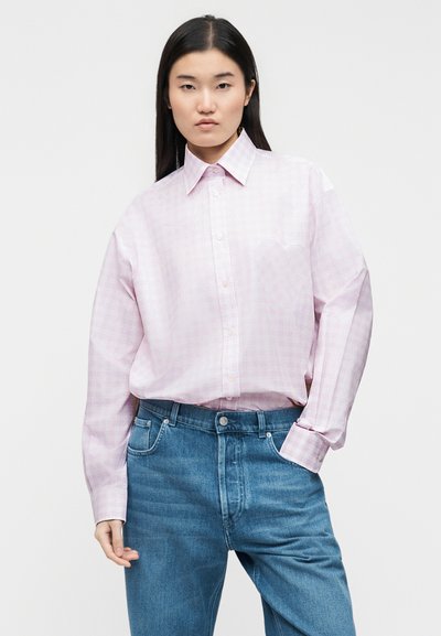 Νεαρή γυναίκα με μακριά σκούρα μαλλιά, φοράει ανοιχτό ροζ πουκάμισο με καρό και μανταλάκια, κλεισμένο μέσα σε μπλε τζιν τζιν, μπροστά σε απλό φόντο.