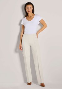 Vrouw staat tegen een lichte muur, gekleed in een witte top met korte mouwen, beige hoge taille broek en bruine puntige hakken.