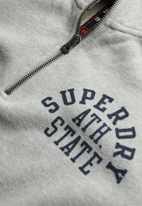 Sudadera con cremallera de color gris claro, confeccionada con una mezcla de tela de algodón, que presenta un estampado en azul marino que dice "SUPERDRY ATH. STATE" y una cremaller metálica.