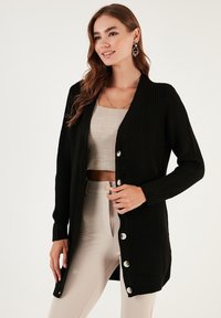 Čierny pletený cardigan s výstrihom do V, dlhými rukávmi a veľkými striebornými gombíkmi vzadu. Nosený cez svetlú blúzku a nohavice.