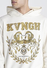 Krémová mikina s kapucí se zlatou grafikou a textem "KVNGH" a heraldickými prvky dvou fénixů mezi dekorativními motivy.