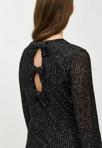 Robe noire à sequins avec un col rond, des manches longues et une découpe dans le dos ornée de deux nœuds. Tissu texturé avec des reflets scintillants.