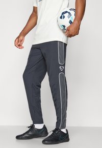 Fekete sportnadrág fehér oldalcsíkokkal és Nike logóval. A modellen foci labda van, fekete sportcipőt visel, melynek türkiz színű részletei vannak.