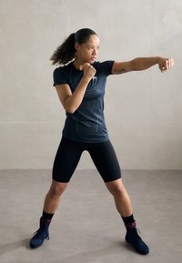 Svart atletisk t-skjorte med kortermet og teksturert mønster, kombinert med svarte shorts. Svart treningste sko med røde detaljer. Nese hevet i en boksestilling.