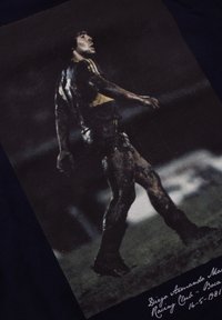 Zwarte t-shirt met een afbeelding van een mannelijke voetballer in een blauw en geel tenue, modderig en in een dynamische pose, met tekst eronder.