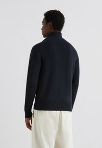 Maglione a coste blu navy con colletto alto, caratterizzato da una superficie testurizzata e dettagli scuri, abbinato a pantaloni chiari.