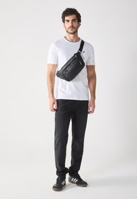 Sacoche noire en bandoulière avec surface texturée, fermeture éclair et sangle réglable, portée sur une chemise blanche et associée à un jean foncé et des chaussures.