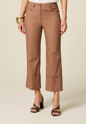 Femme portant un pantalon ample beige coupé court, un haut blanc, des sandales à talons foncées et des bracelets dorés, debout devant un fond uni.