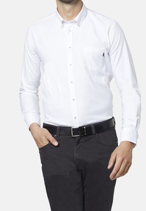 Mężczyzna w dopasowanej białej koszuli z guzikami i kieszenią na piersi, czarnym pasku i ciemnych spodniach, z jedną ręką w kieszeni.