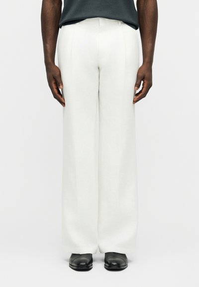 Homme portant un pantalon large blanc, des chaussures noires et une chemise foncée rentrée, debout devant un fond clair uni.