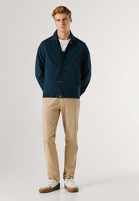 Chaqueta ligera azul marino con cierre de botones, usada sobre una camiseta blanca. Combinada con pantalones chinos beige y zapatillas blancas. Textura suave.