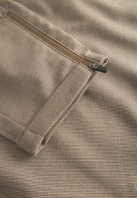 Bežasti tekstil s grubom teksturom, s džepom na zatvarač. Zatvarač je metalik s crnim drškom, integriran u tkaninu.