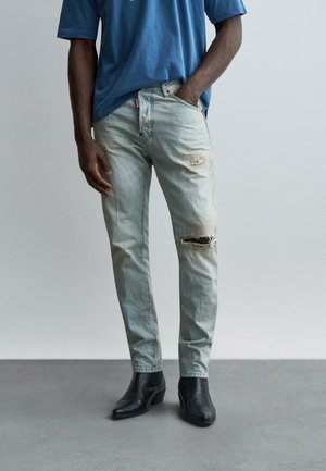 Mann trägt hellblaue, zerrissene Jeans, eine Hand in der Tasche, kombiniert mit schwarzen Stiefeletten auf grauem Boden vor einem schlichten Hintergrund.