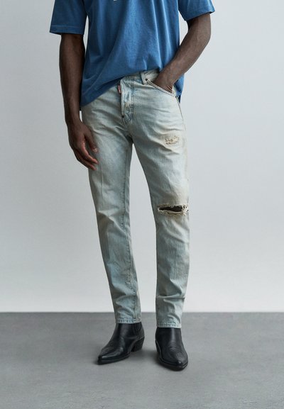Homme portant un jean déchiré bleu clair, une main dans la poche, associé à des bottines noires sur un sol gris devant un fond uni.