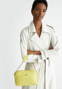 Borsa a tracolla gialla trapuntata con catena e hardware color oro, caratterizzata da una texture liscia e un dettaglio con logo. Abbinata a un cappotto bianco.