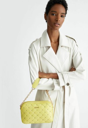 Borsa a tracolla gialla trapuntata con catena e hardware color oro, caratterizzata da una texture liscia e un dettaglio con logo. Abbinata a un cappotto bianco.