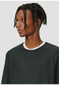 Donkergrijs longsleeve shirt met een ronde hals en een witte binnenkraag, gemaakt van een textuurstof, gepresenteerd op een persoon die naar rechts kijkt.